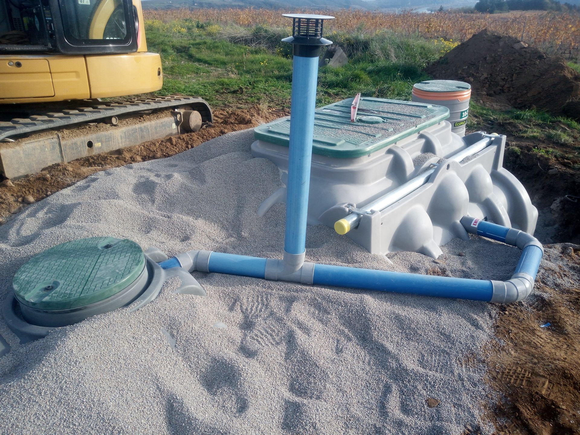 Site d'installation d'une fosse septique avec cuve, tuyaux et pelle mécanique sur un lit de gravier.