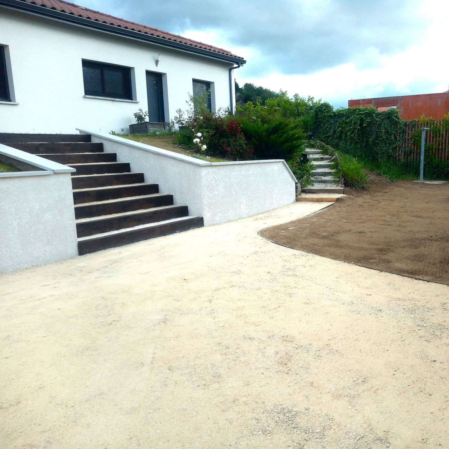 Maison avec escalier, allée et jardin sous un ciel partiellement nuageux.