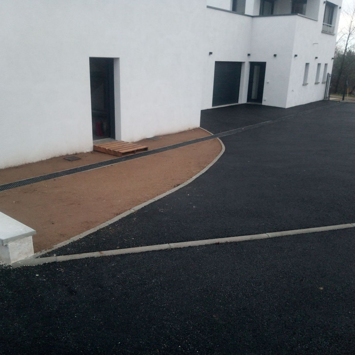 Allée en asphalte noir jouxtant un bâtiment blanc. Un chemin beige sépare l'allée du bâtiment.
