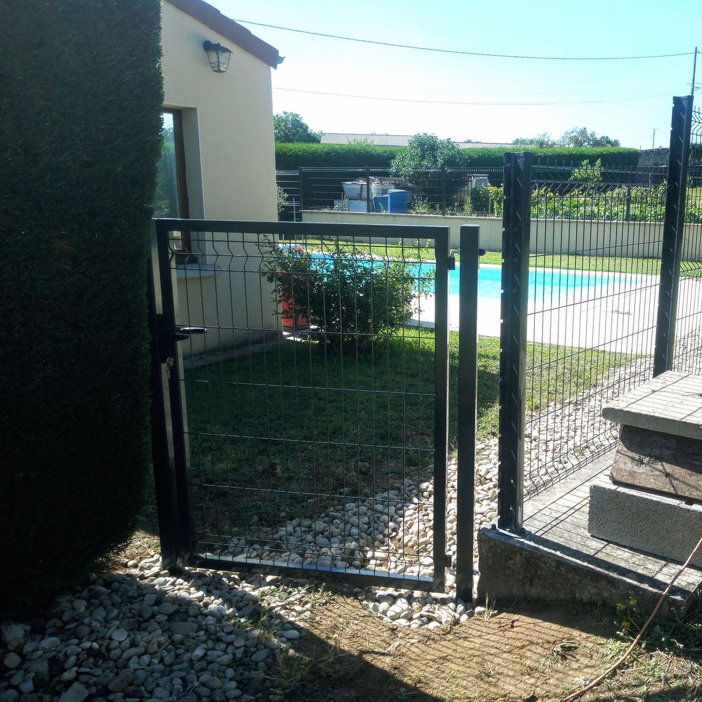 Portail en métal noir avec grillage, donnant sur un jardin avec piscine, pelouse et maison beige.