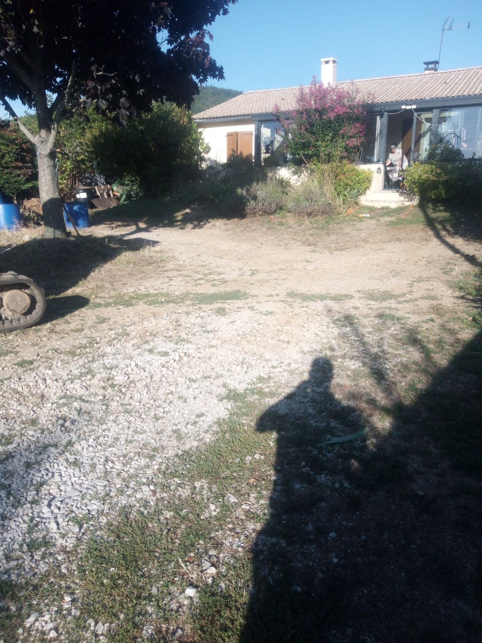 Une cour ensoleillée, recouverte de gravier et d'herbe, mène à une maison avec un porche. Une ombre se projette sur le sol.