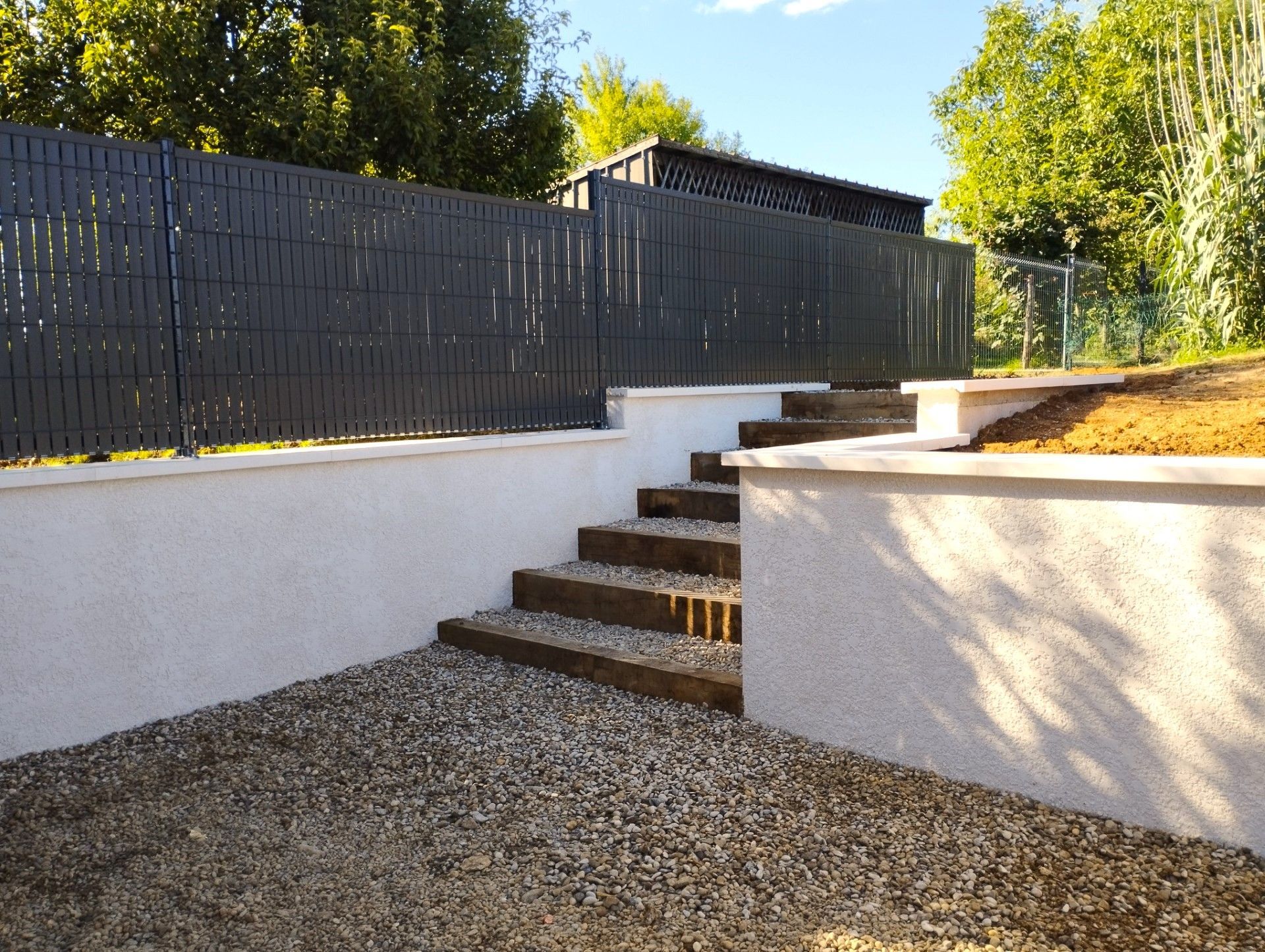 Clôture grise sur un mur de soutènement blanc, avec des marches en gravier et en pierre menant à l'étage.