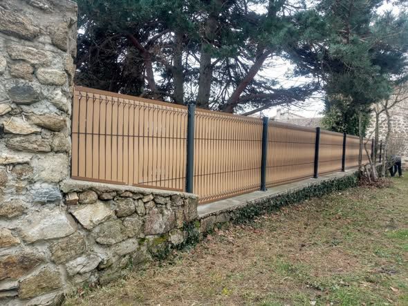 Mur en pierre avec clôture en bambou beige et poteaux noirs dans une zone herbeuse. Arbres en arrière-plan.