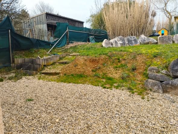 Une zone de gravier en pente avec des marches et un monticule de terre, menant à une structure ombragée et verdoyante et à un fourré de bambous.