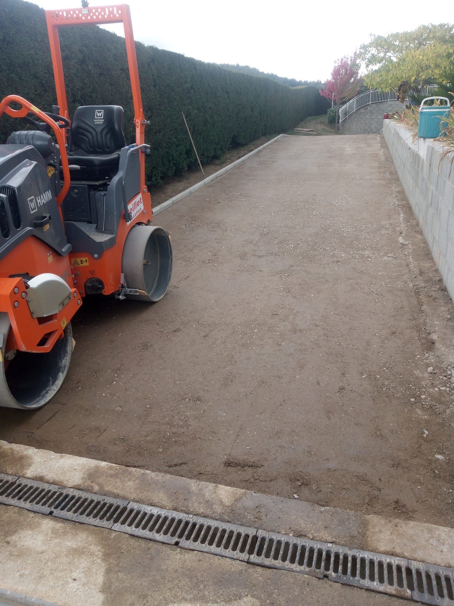 Un rouleau compresseur compacte du gravier sur une allée. Une machine orange sur une surface grise, avec une haie en arrière-plan.