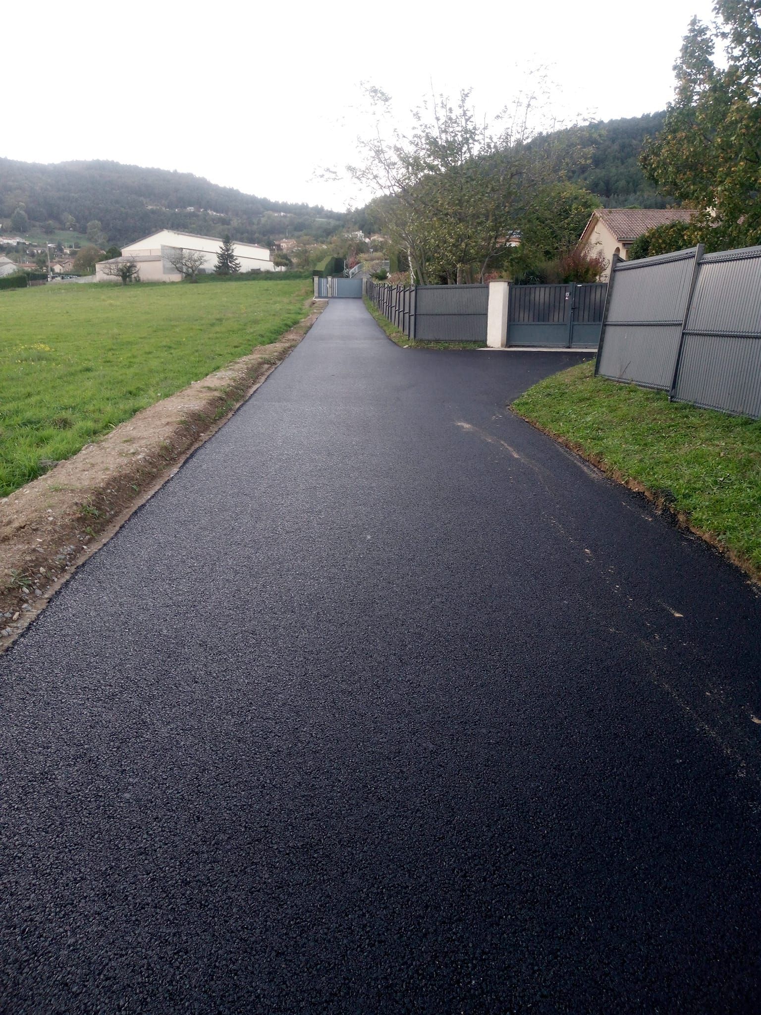 Chemin d'asphalte noir longeant l'herbe et une clôture, menant vers les maisons et les collines.