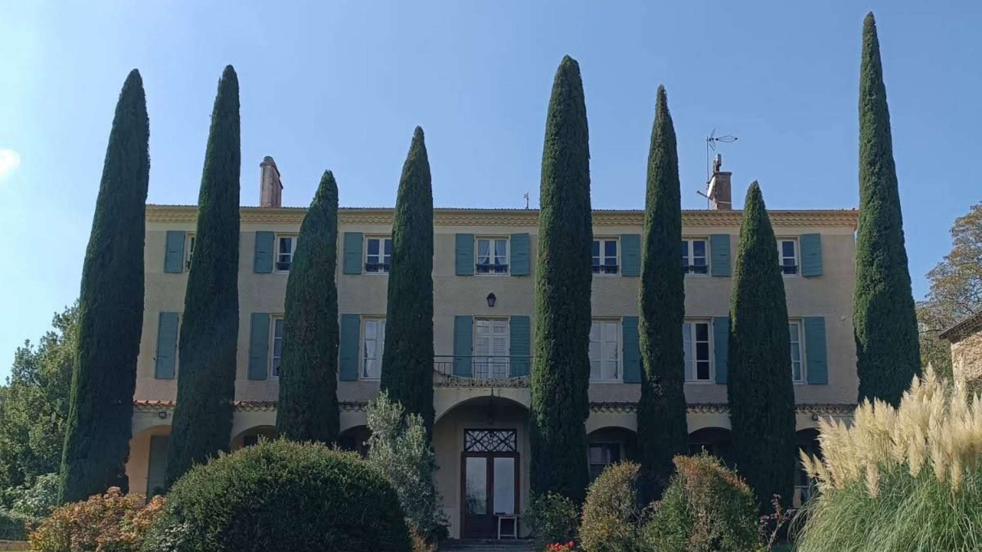 Un imposant bâtiment beige aux volets verts, encadré par de grands arbres élancés se détachant sur un ciel d'un bleu limpide.