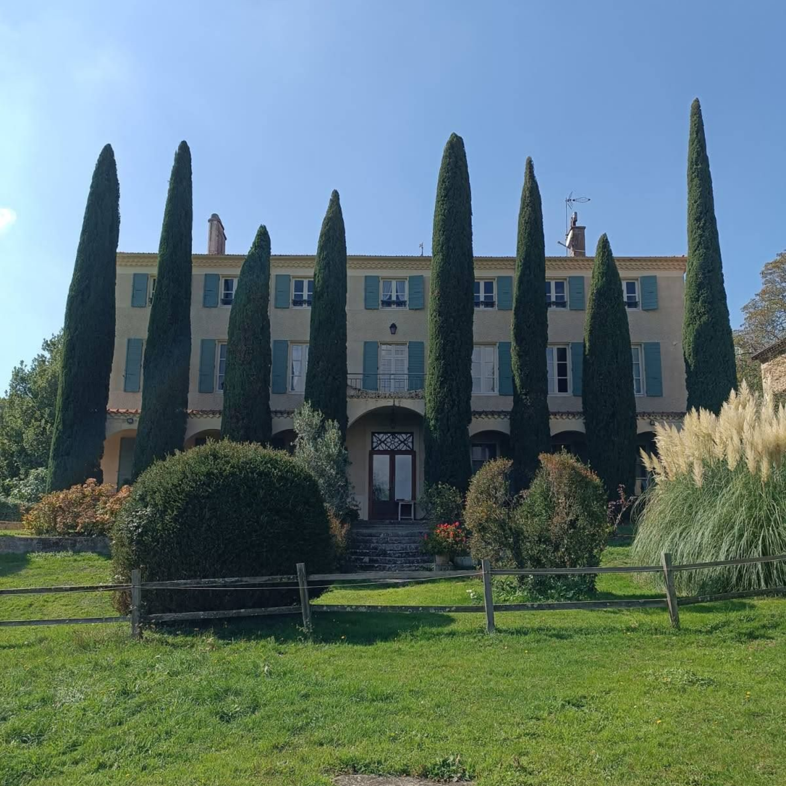 Grand bâtiment beige avec de grands cyprès à l'avant, pelouse verte, volets bleus et ciel ensoleillé.