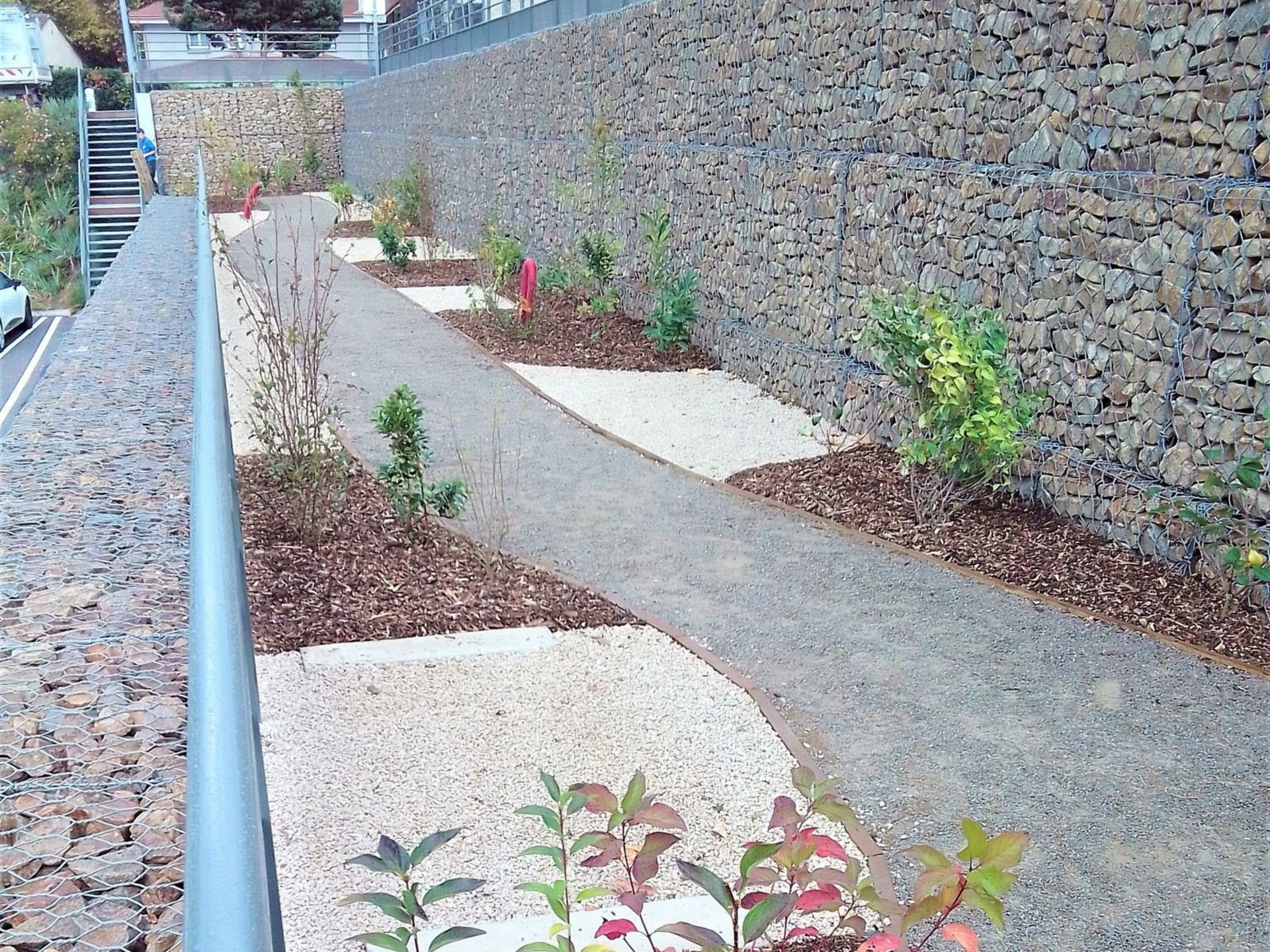 Chemin longeant un mur de soutènement bordé de parterres fleuris. Mur en pierre à droite, rambarde à gauche.