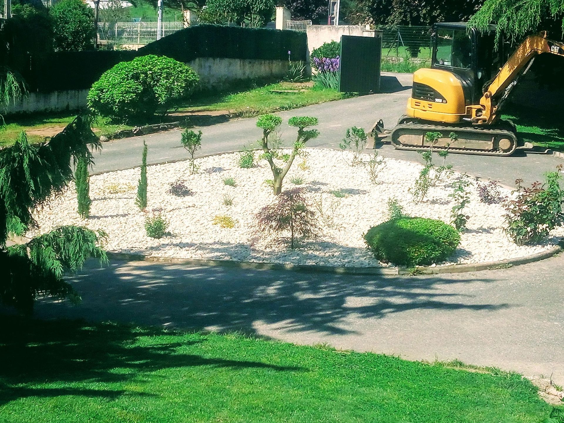 Une petite pelleteuse travaille dans un parterre de jardin rond rempli de pierres blanches et de plantes diverses ; une route et des arbres entourent le tout.