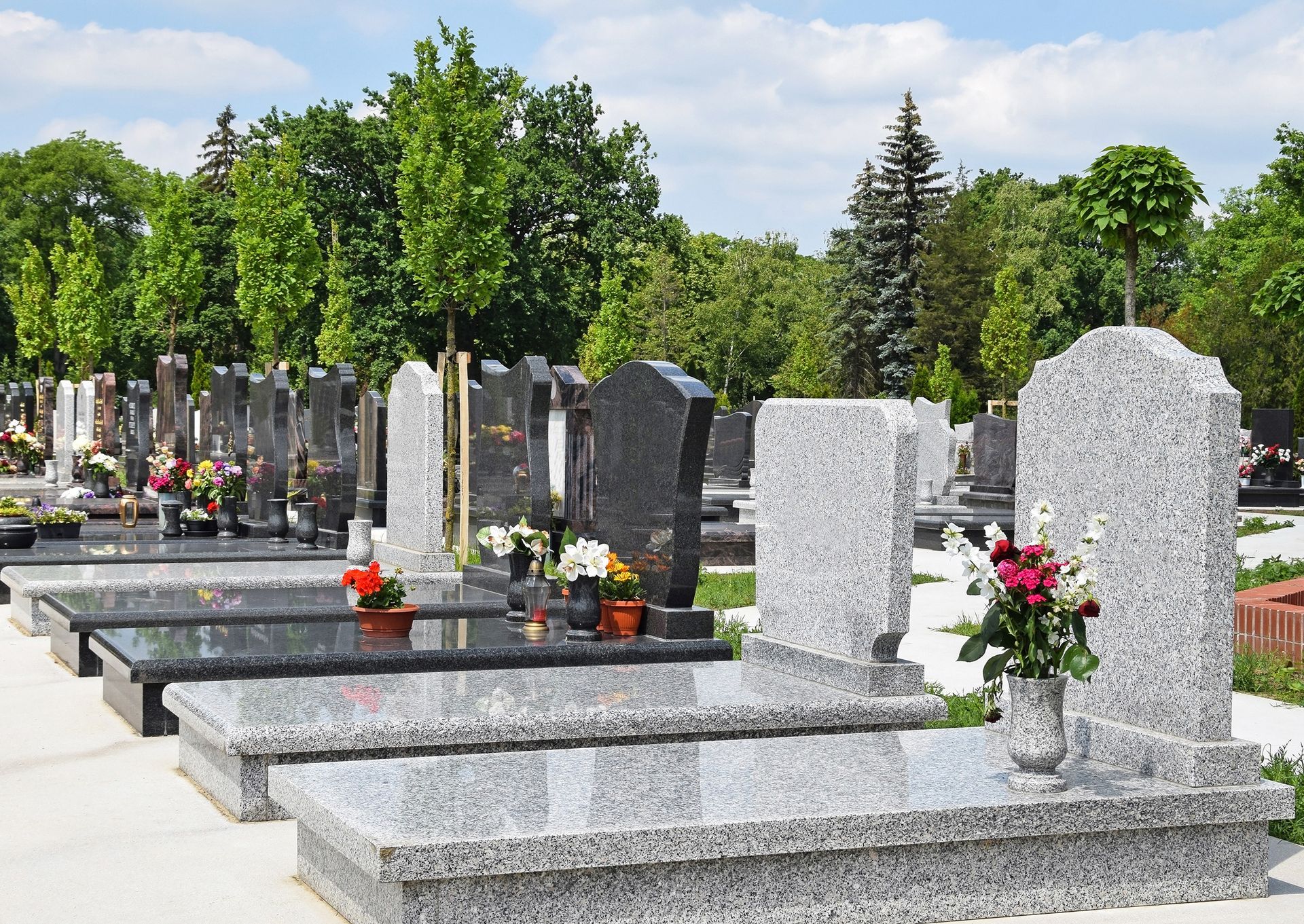 Des rangées de pierres tombales avec des fleurs dans un cimetière, et des arbres verts en arrière-plan.