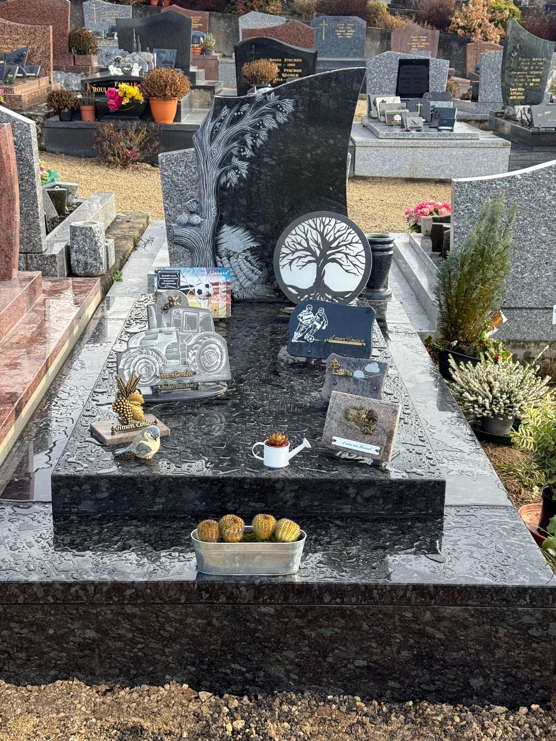 Une pierre tombale moderne en granit noir décorée de fleurs et de petits ornements, dans un cimetière.