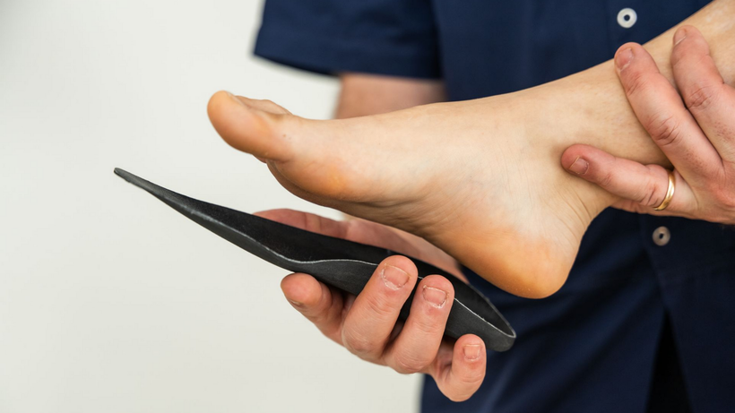 Pied tenu par une personne portant une chemise bleue et une semelle intérieure noire ; fond clair.