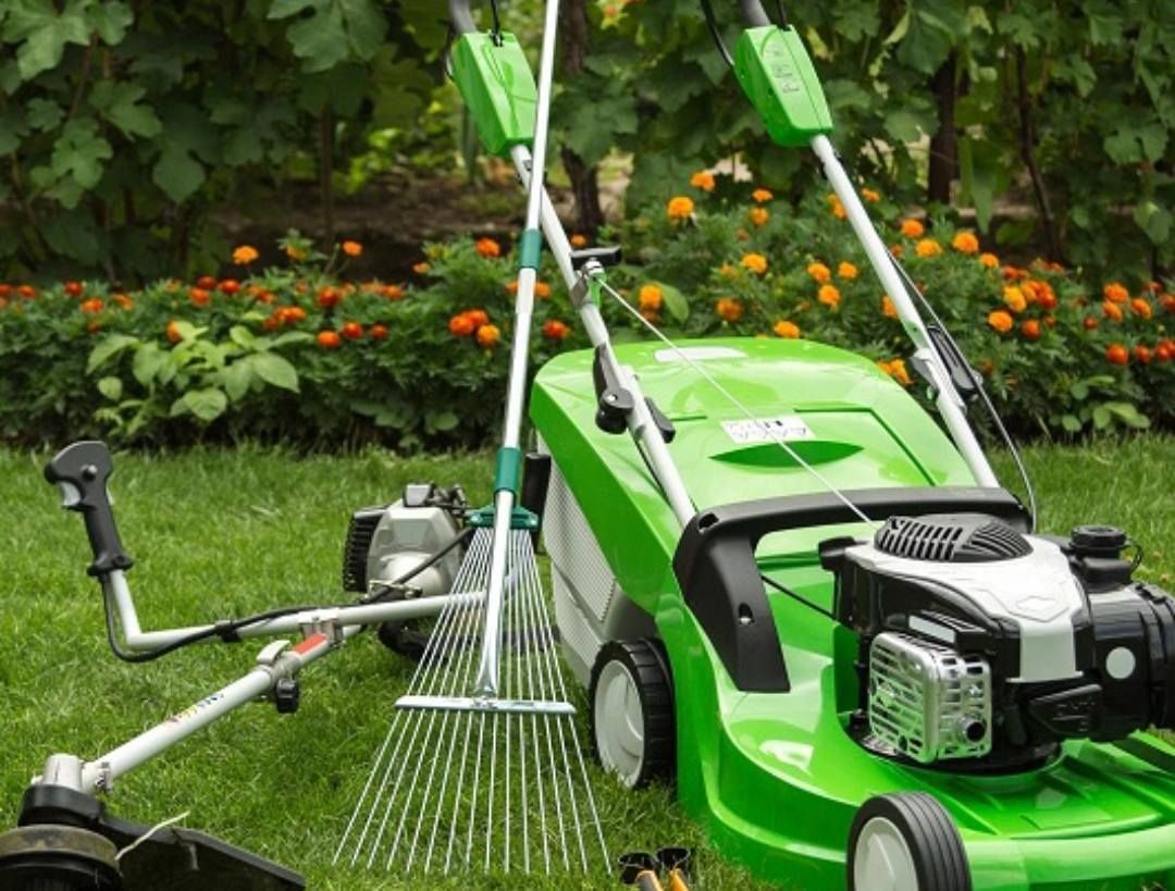 Tondeuse à gazon verte, coupe-bordures et râteau sur l'herbe devant les fleurs.
