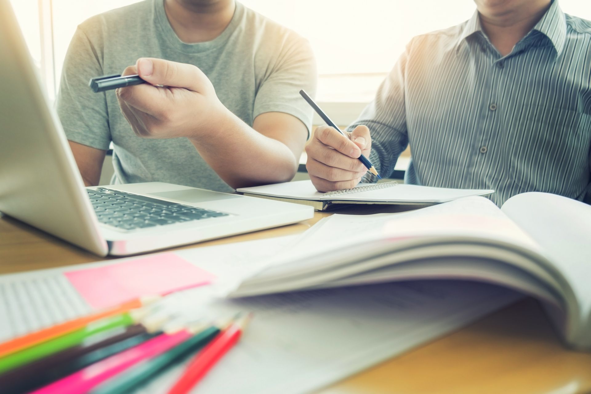 Deux personnes travaillent à un bureau avec un ordinateur portable, des cahiers et des crayons de couleur. L'une d'elles pointe un stylo.