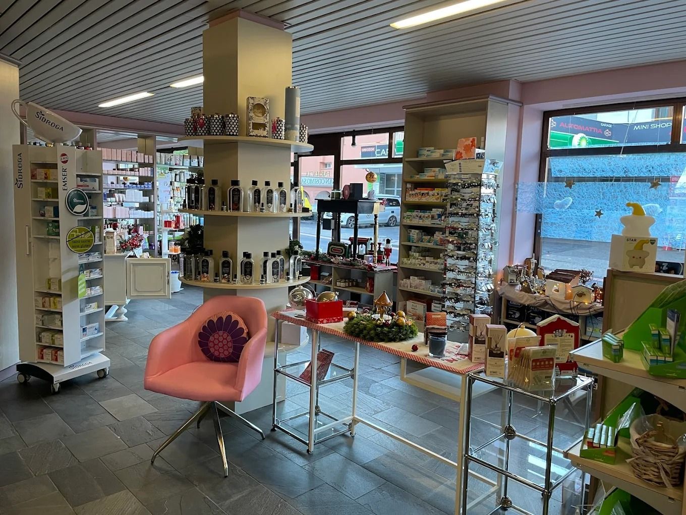 Luminoso interno di una boutique con scaffali pieni di prodotti, tavoli espositivi e una sedia rosa vicino alle grandi finestre.