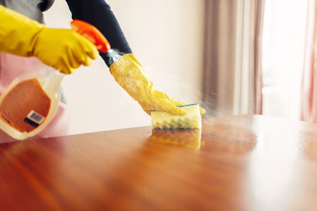 Una persona que lleva guantes amarillos rocía un limpiador sobre una mesa de madera y la limpia con una esponja.