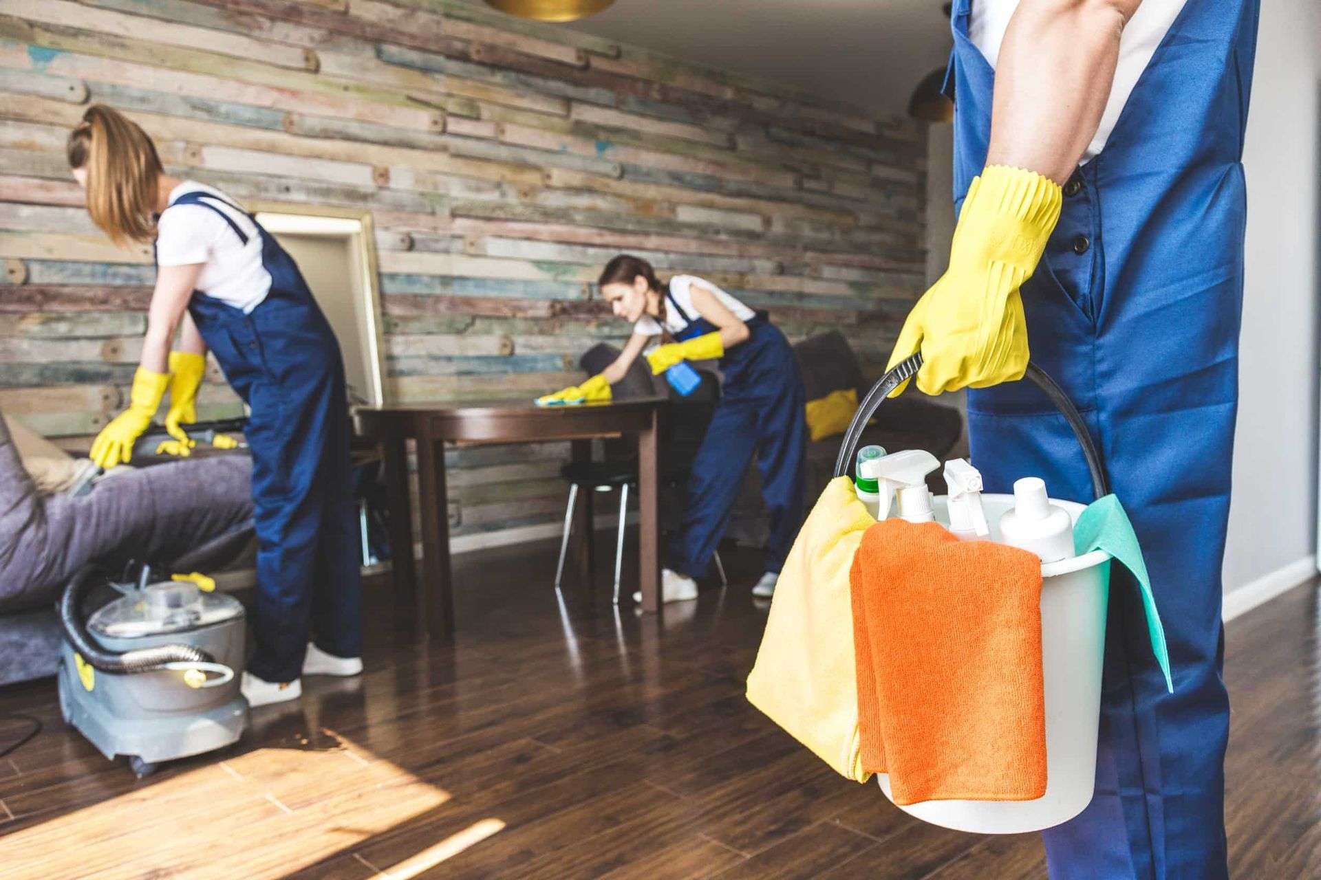Tres personas con monos azules limpiando una habitación, una de ellas sosteniendo un balde de limpieza con suministros.