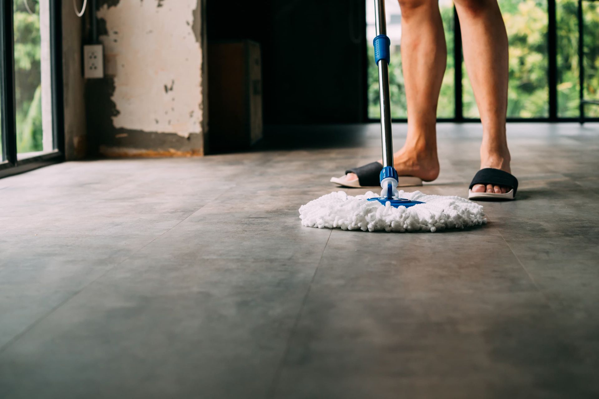 Persona trapeando un piso gris con un trapeador azul y blanco, usando sandalias en una habitación con grandes ventanas.