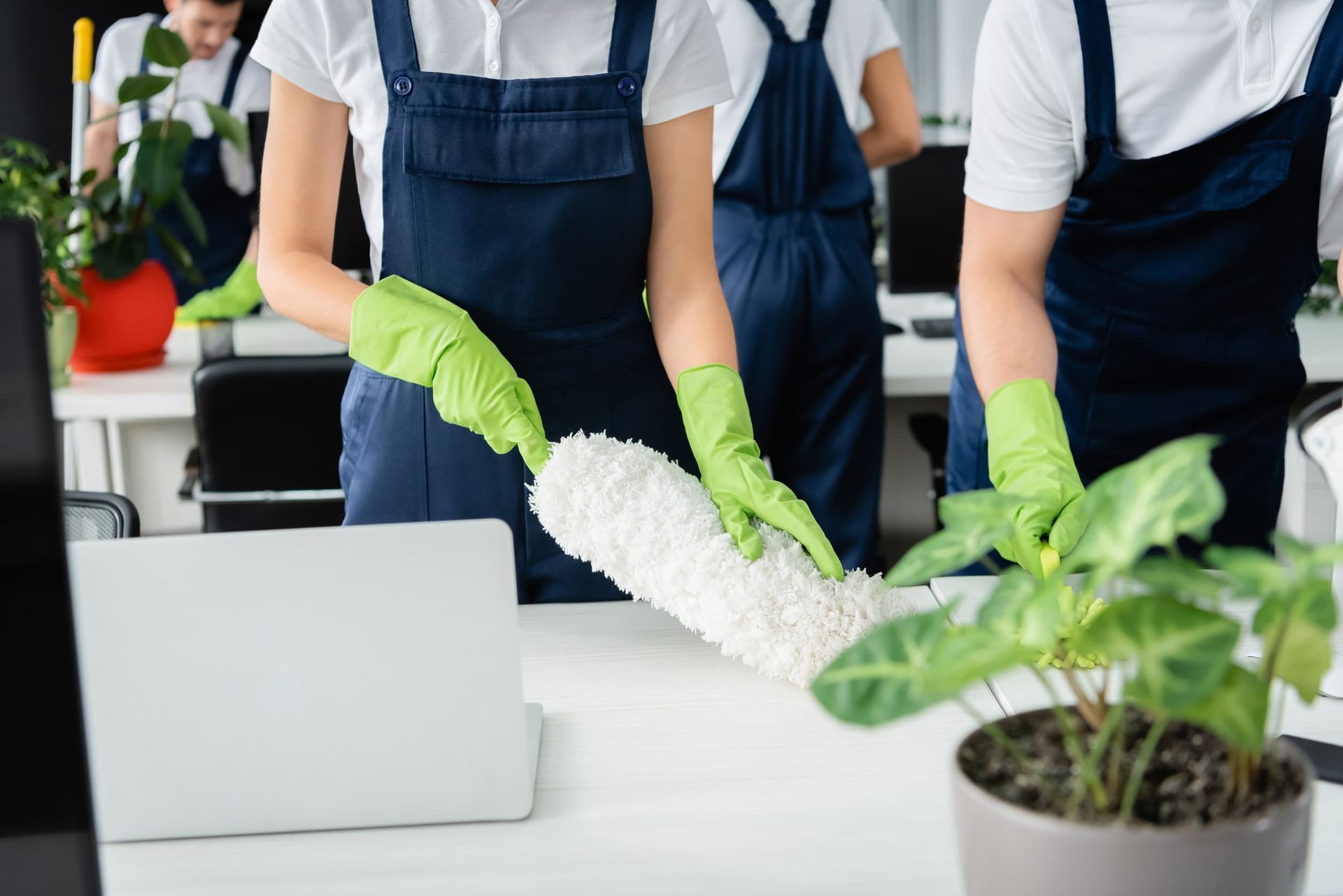 Personal de limpieza de oficinas quitando el polvo de los escritorios; vistiendo overoles azules y guantes verdes.