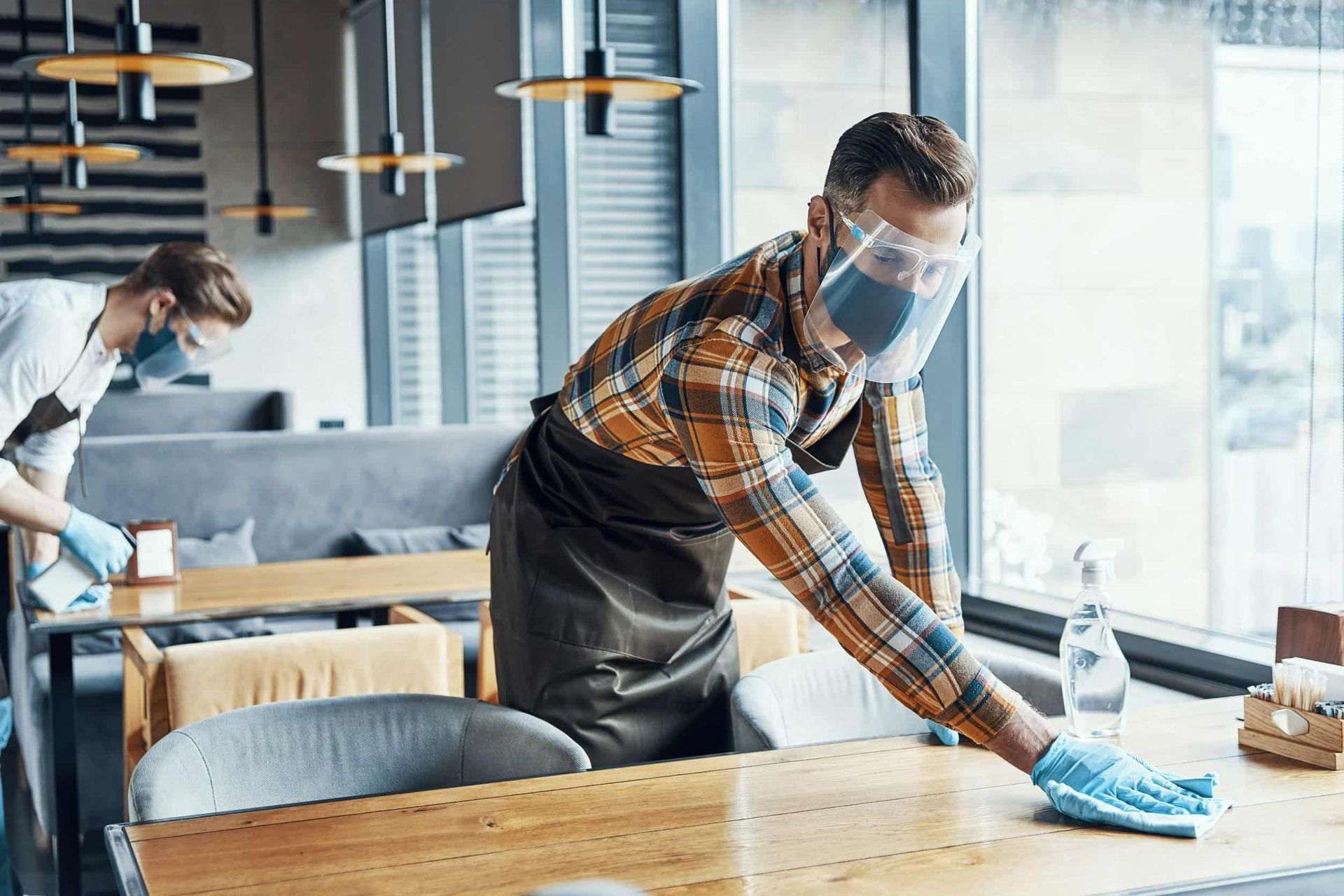 Trabajadores de restaurantes limpiando mesas, usando mascarillas y protectores faciales.