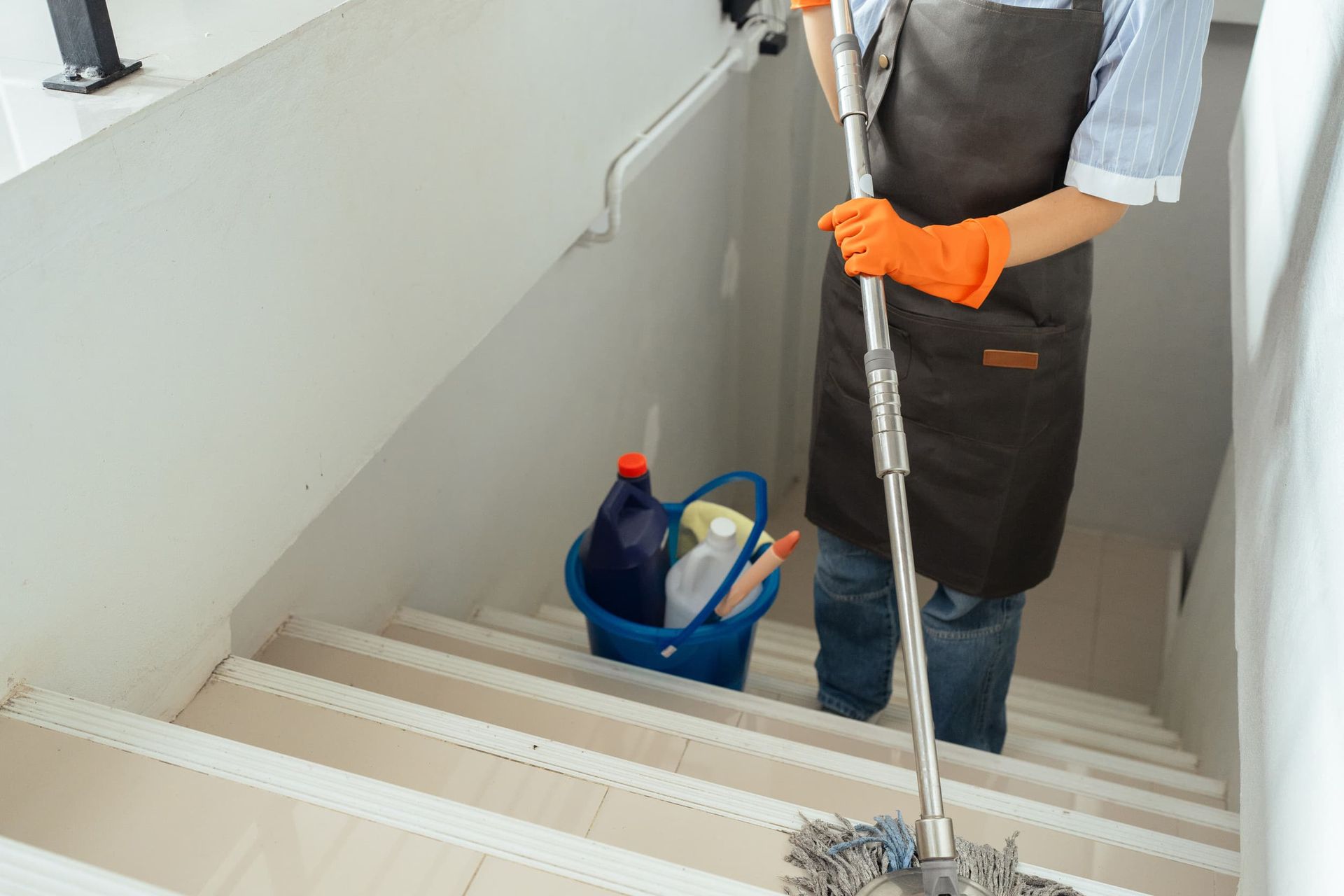 Persona limpiando escaleras con productos de limpieza en un balde cercano; viste delantal y guantes naranjas.