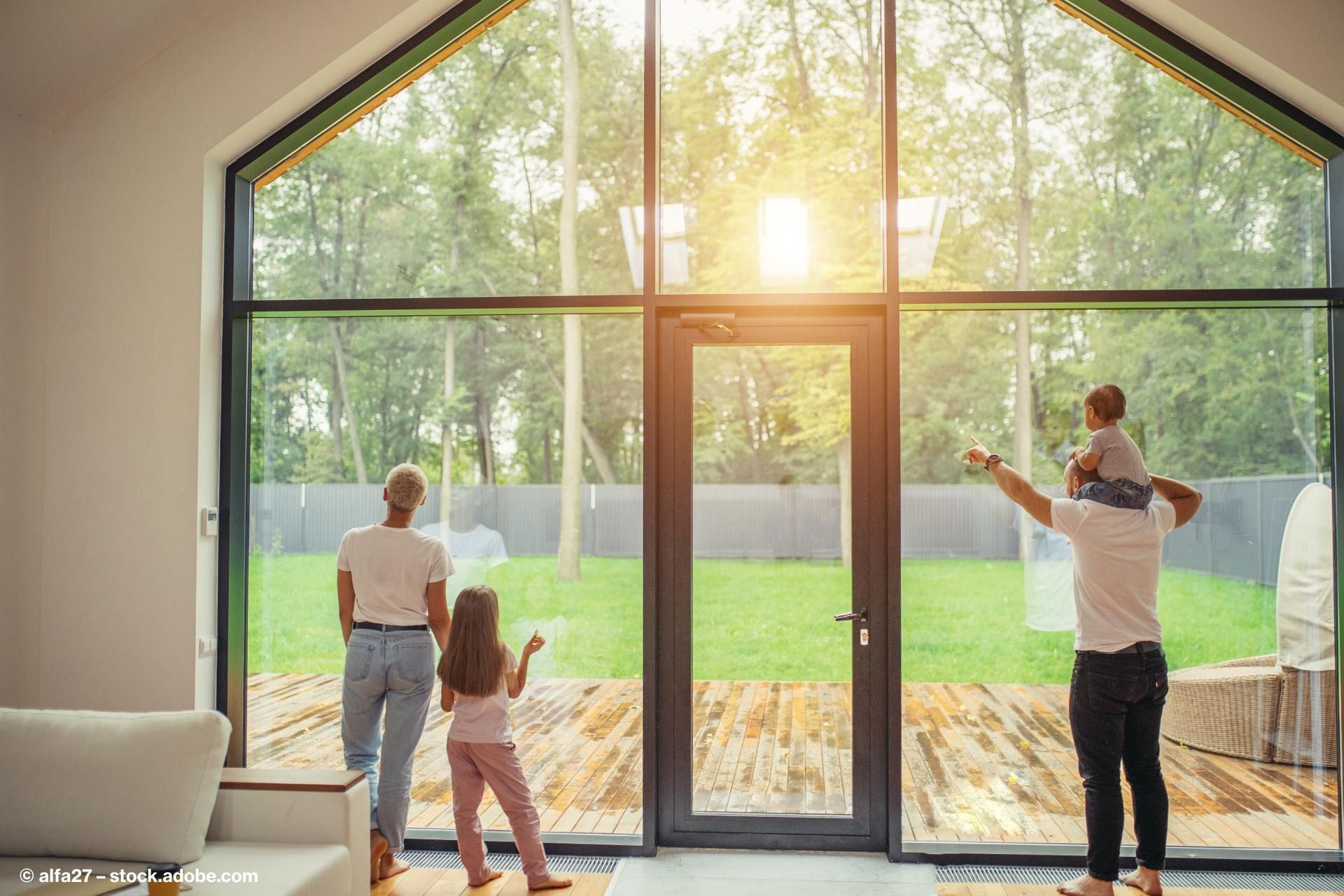 eine vierköpfige Familie steht vor einer großen Fensterfront und schaut in den Garten