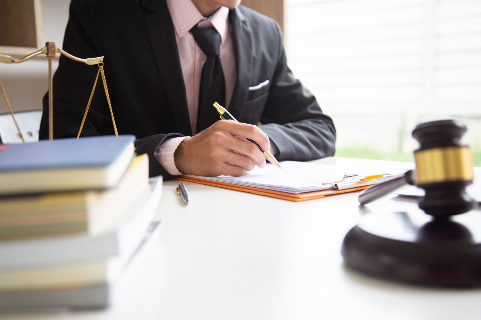 Abogado con traje negro escribiendo en un escritorio, con balanza de justicia, mazo y libros visibles.
