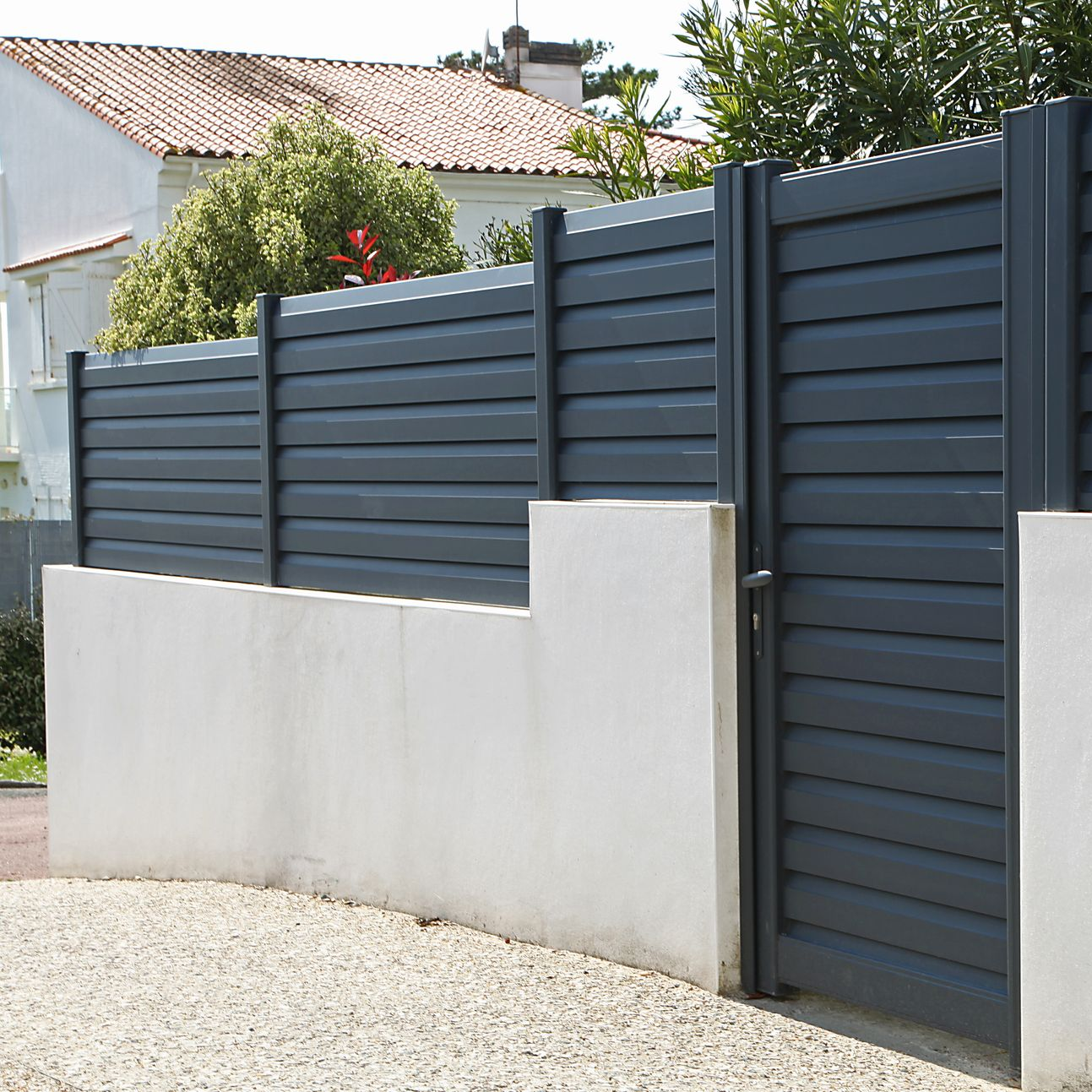 Clôture foncée au-dessus d'un mur en stuc blanc, à côté d'une allée de gravier.
