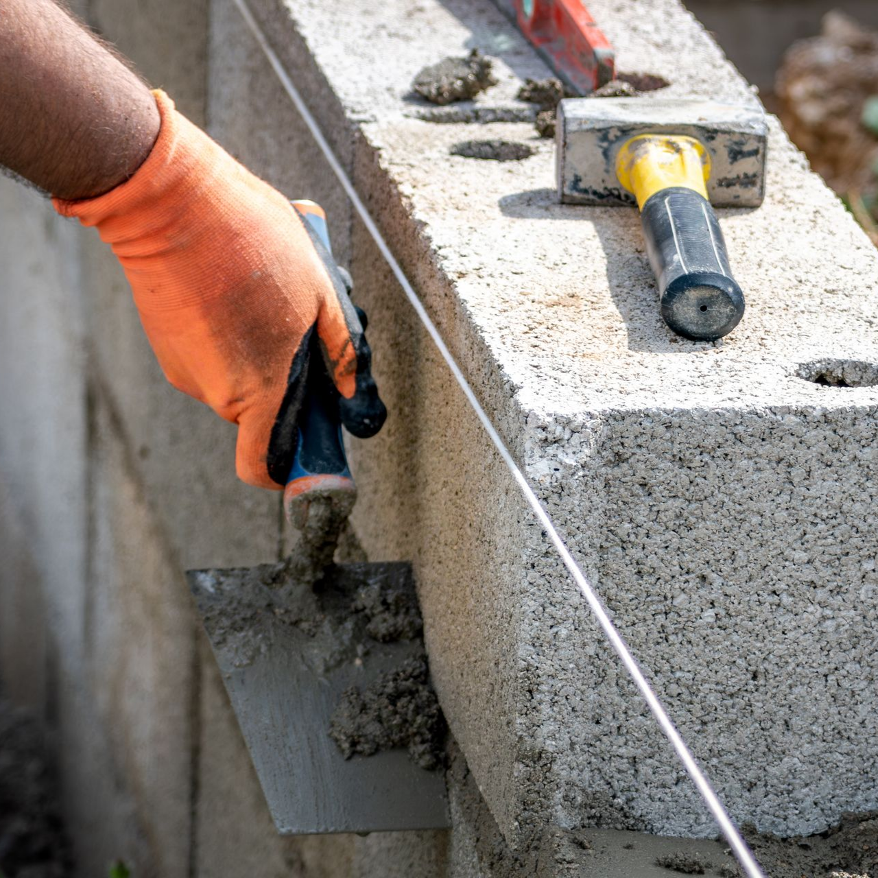 Main gantée appliquant du mortier sur un mur en parpaings à l'aide d'une truelle et d'un niveau.