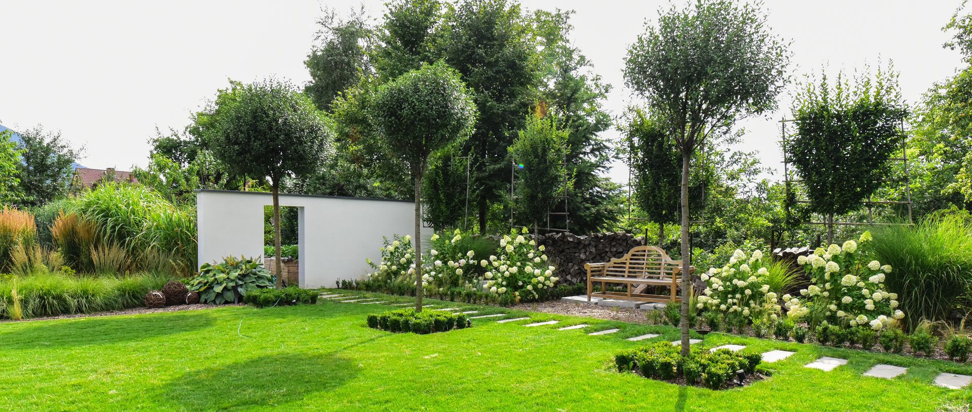 Jardin verdoyant avec arbres, arbustes, un mur blanc et un petit banc.