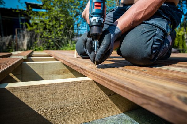 Une personne utilise une perceuse électrique pour fixer une planche de terrasse en bois à l'extérieur.
