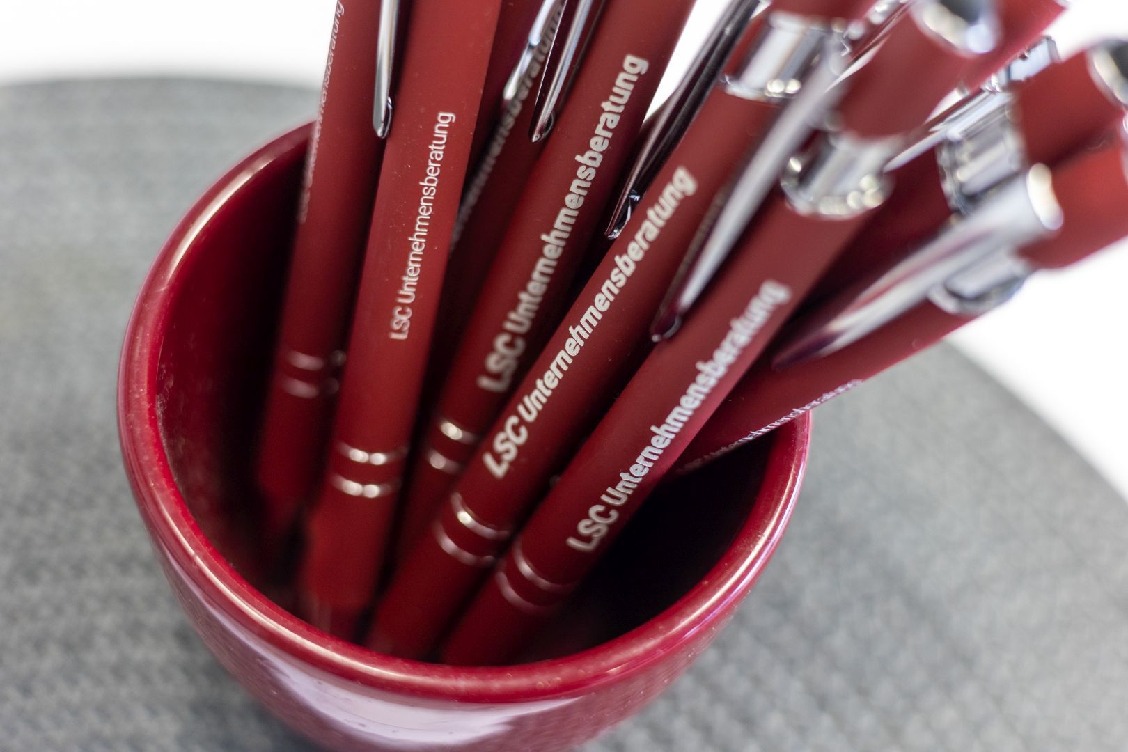 Eine rote Keramiktasse, gefüllt mit roten Stiften, auf grauer Oberfläche