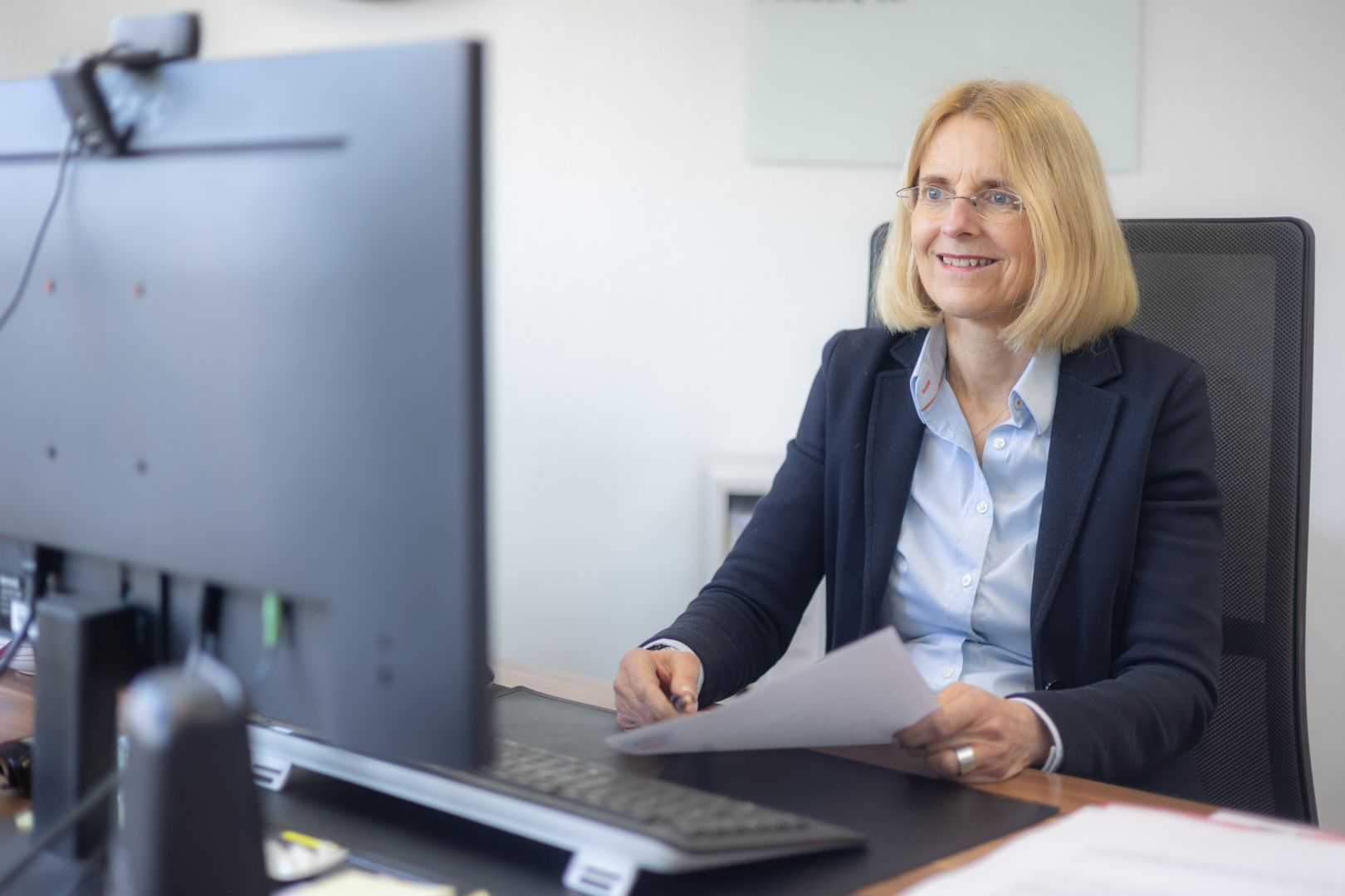 Frau im Blazer lächelt an einem Schreibtisch mit Computer und Papieren in der Hand in einem Büro.