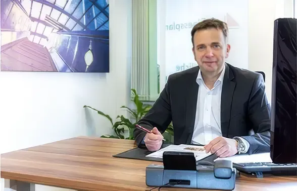 Ein Mann im Anzug sitzt an einem Schreibtisch in einem Büro, er hält einen Stift in der Hand, neben ihm stehen Papiere und ein Laptop.