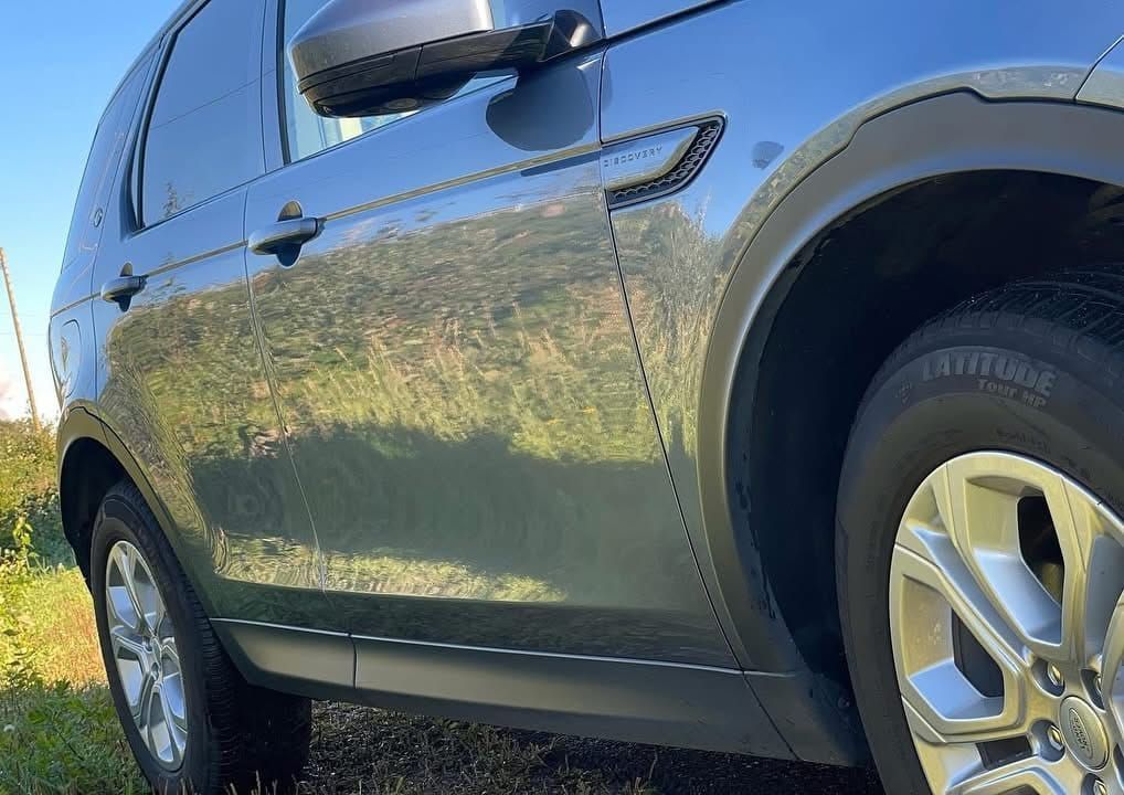 Lähikuva tummasta nurmikolle pysäköidystä katumaasturista, jonka auringonvalo heijastuu sivusta ja renkaista.
