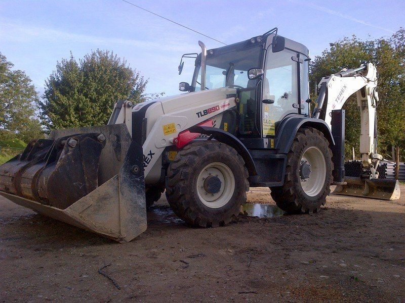 Une chargeuse-pelleteuse Terex blanche et rouge est stationnée sur une surface de gravier, avec le godet et le bras de terrassement déployés.