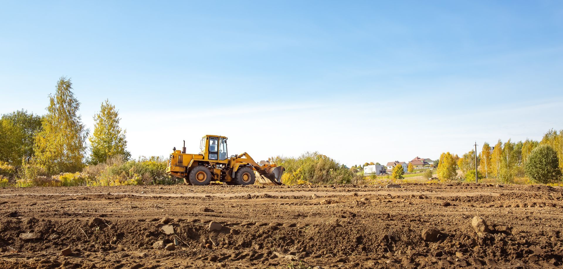 Un bulldozer jaune laboure un champ sous un ciel bleu dégagé, avec des arbres en arrière-plan.