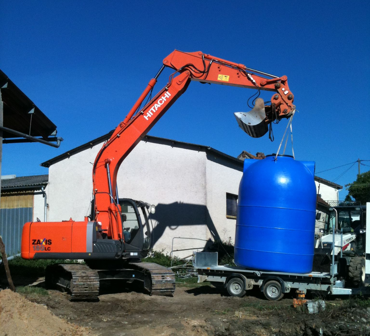 Une pelle mécanique Hitachi orange soulève un grand réservoir cylindrique bleu d'une remorque .