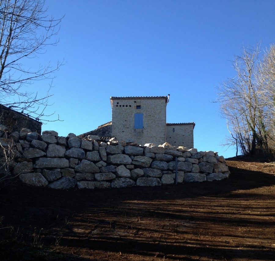 Bâtiment en pierre aux volets bleus, derrière un grand mur de soutènement en pierre, sous un ciel d'un bleu limpide.