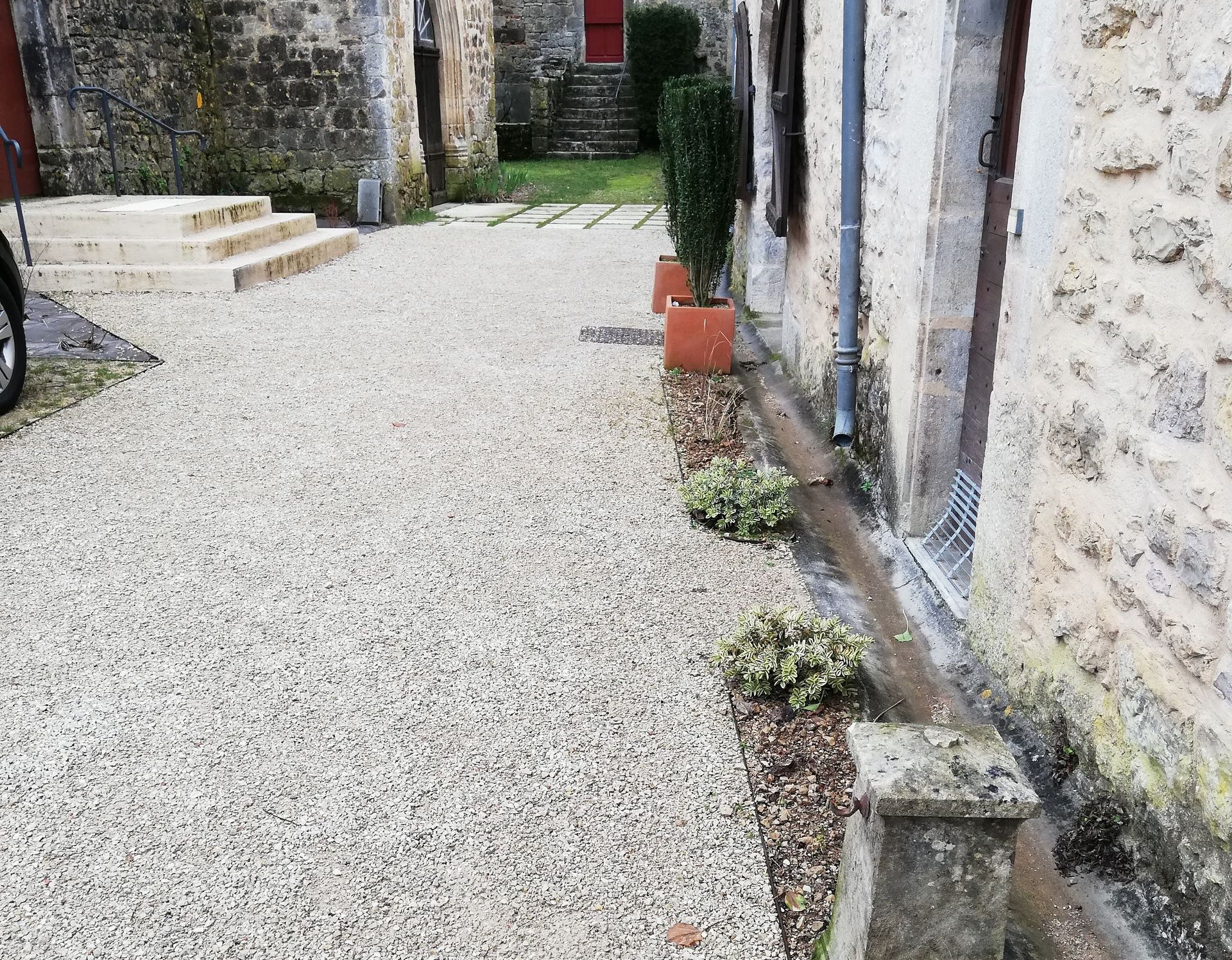 Allée de gravier menant à des marches en pierre et aux portes des bâtiments. Arbustes et plantes en pot sont visibles.