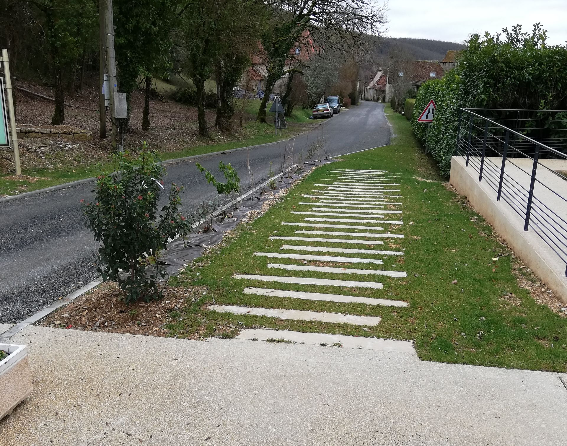 Scène de rue : route asphaltée, chemin de pierre, herbe verte, buissons, maisons et ciel nuageux.