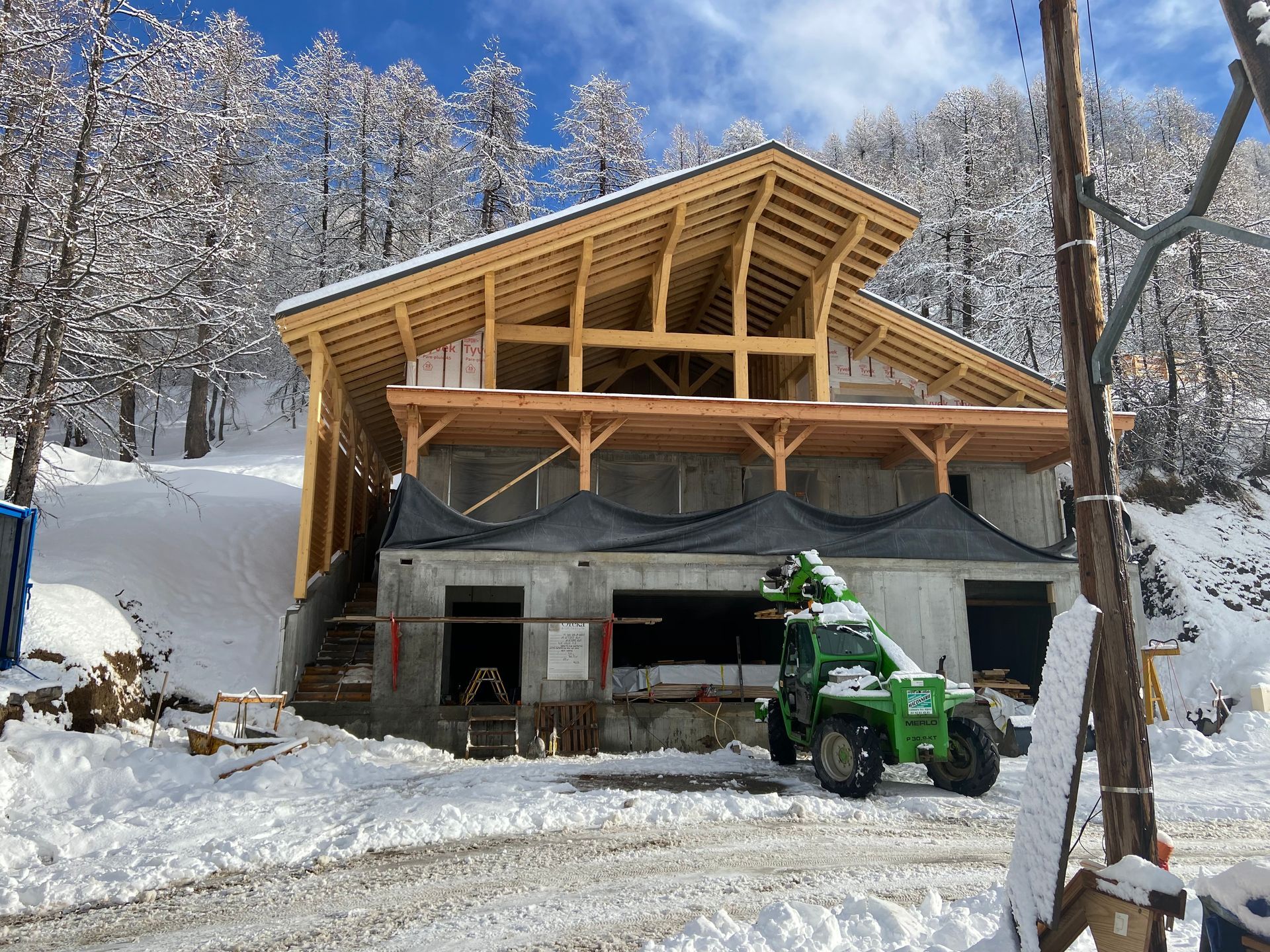 Dans un paysage montagneux enneigé, une structure en bois partiellement construite repose sur des fondations en béton.
