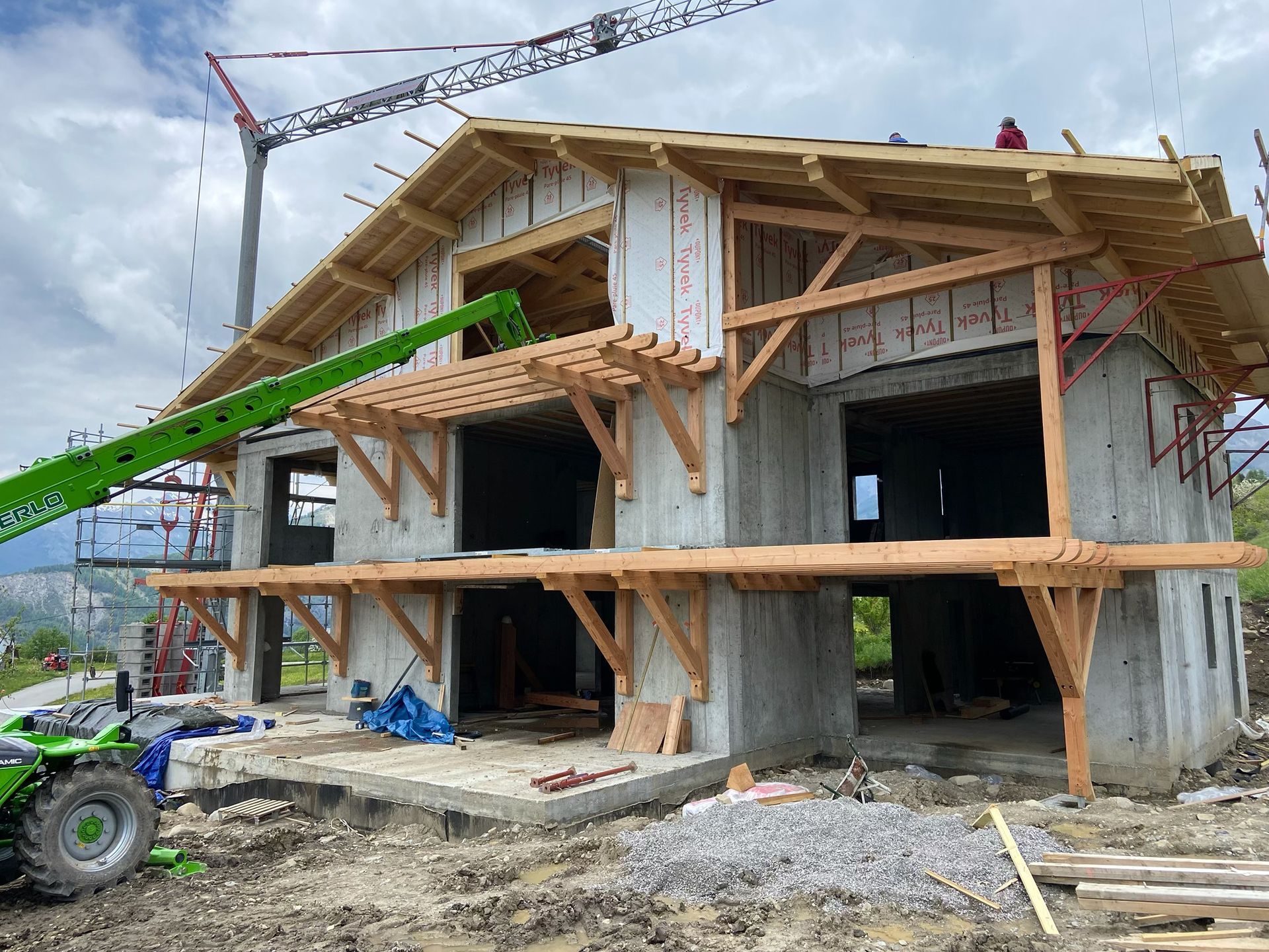 Maison en construction avec une ossature en bois, des piliers en béton et un bras de grue vert sur la gauche.