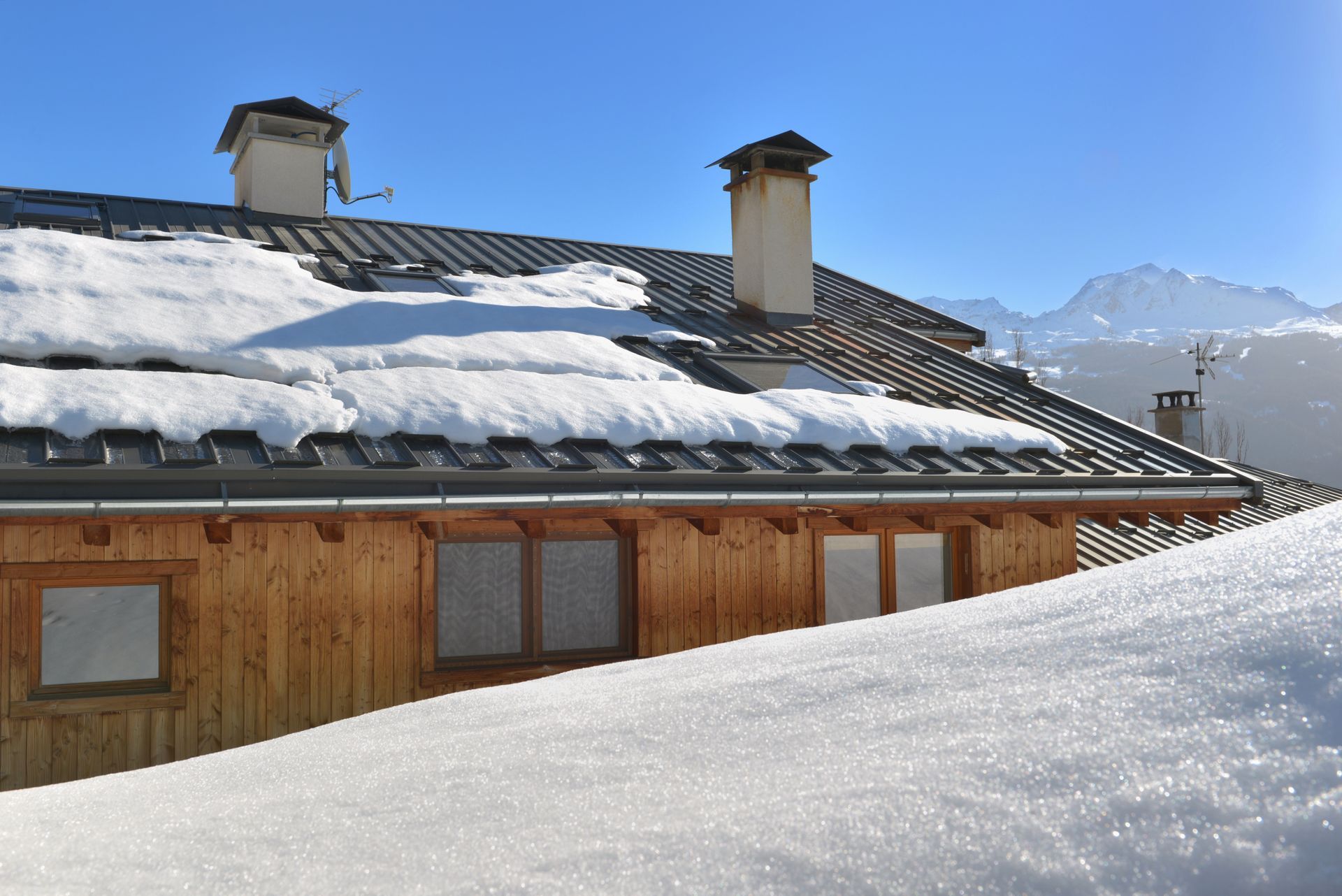 Une cabane en bois au toit et aux cheminées enneigés se détachant sur un ciel bleu et des montagnes enneigées.