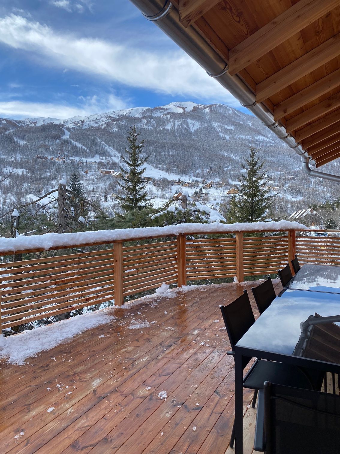 Vue sur les montagnes enneigées depuis une terrasse en bois avec rambarde et table sous un avant-toit