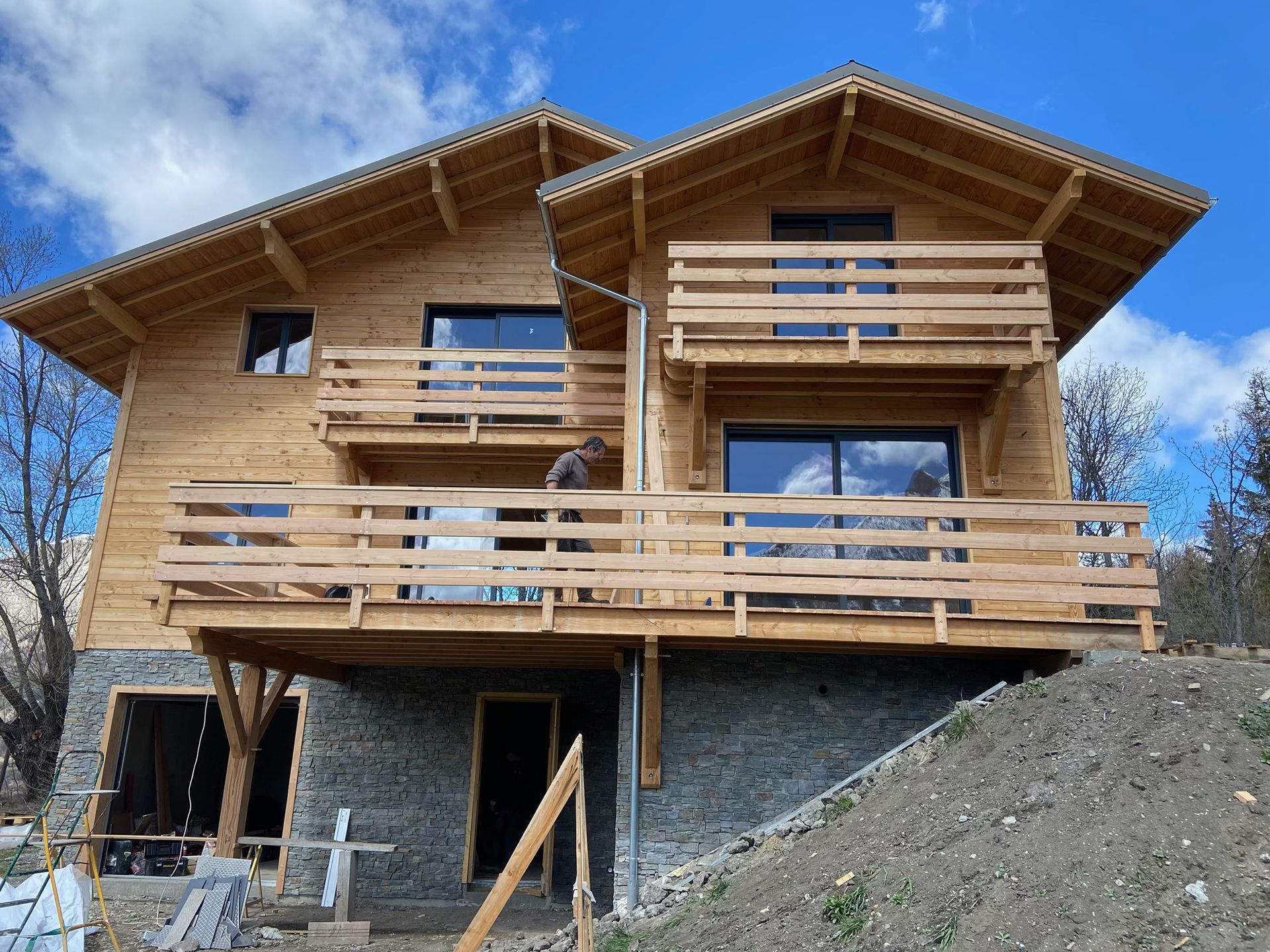 Maison en bois en construction à flanc de colline, avec balcons et fondations en pierre apparente.