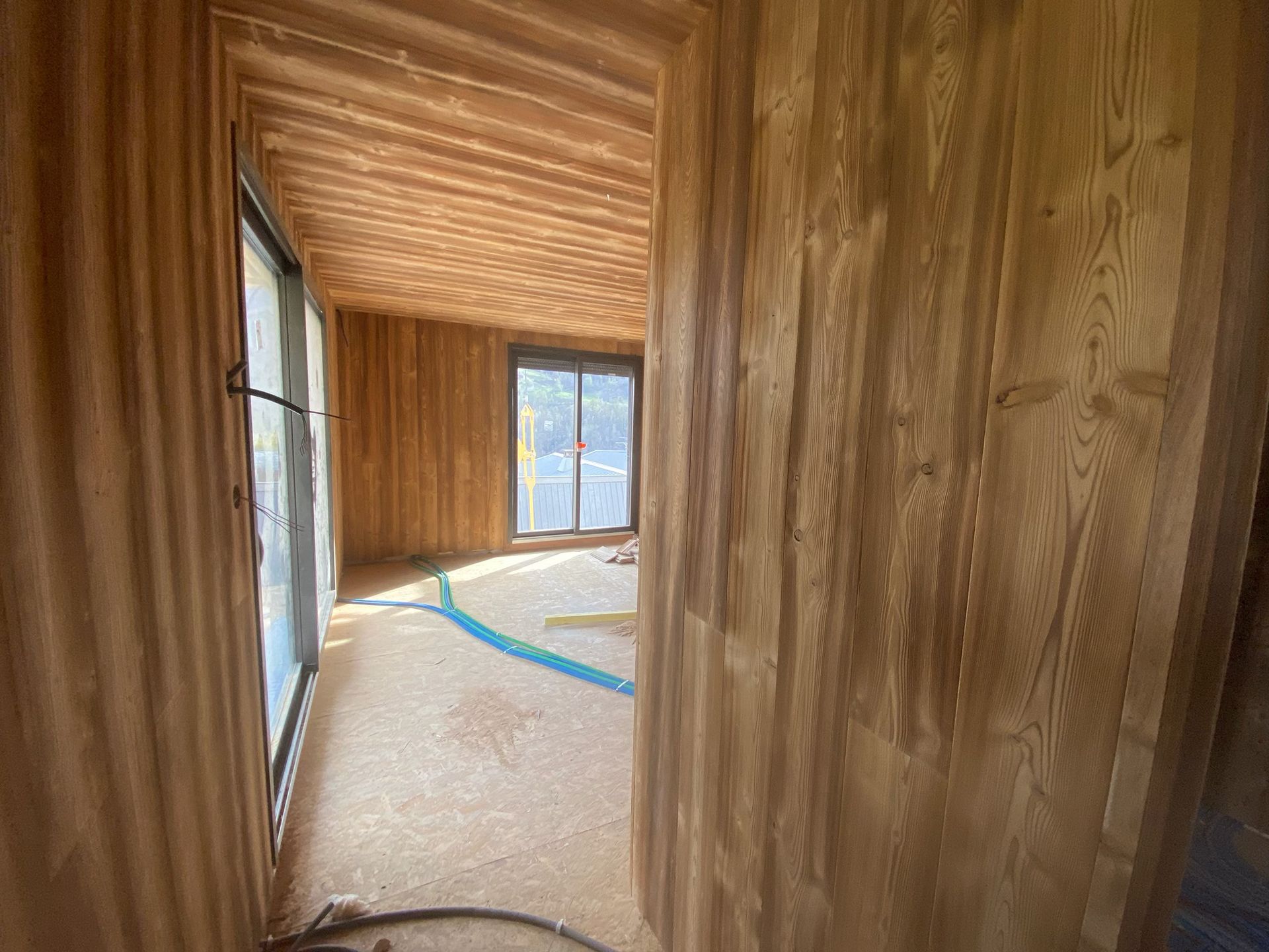 Vue intérieure d'une pièce en construction, avec des murs lambrissés, un sous-plancher et de grandes fenêtres.