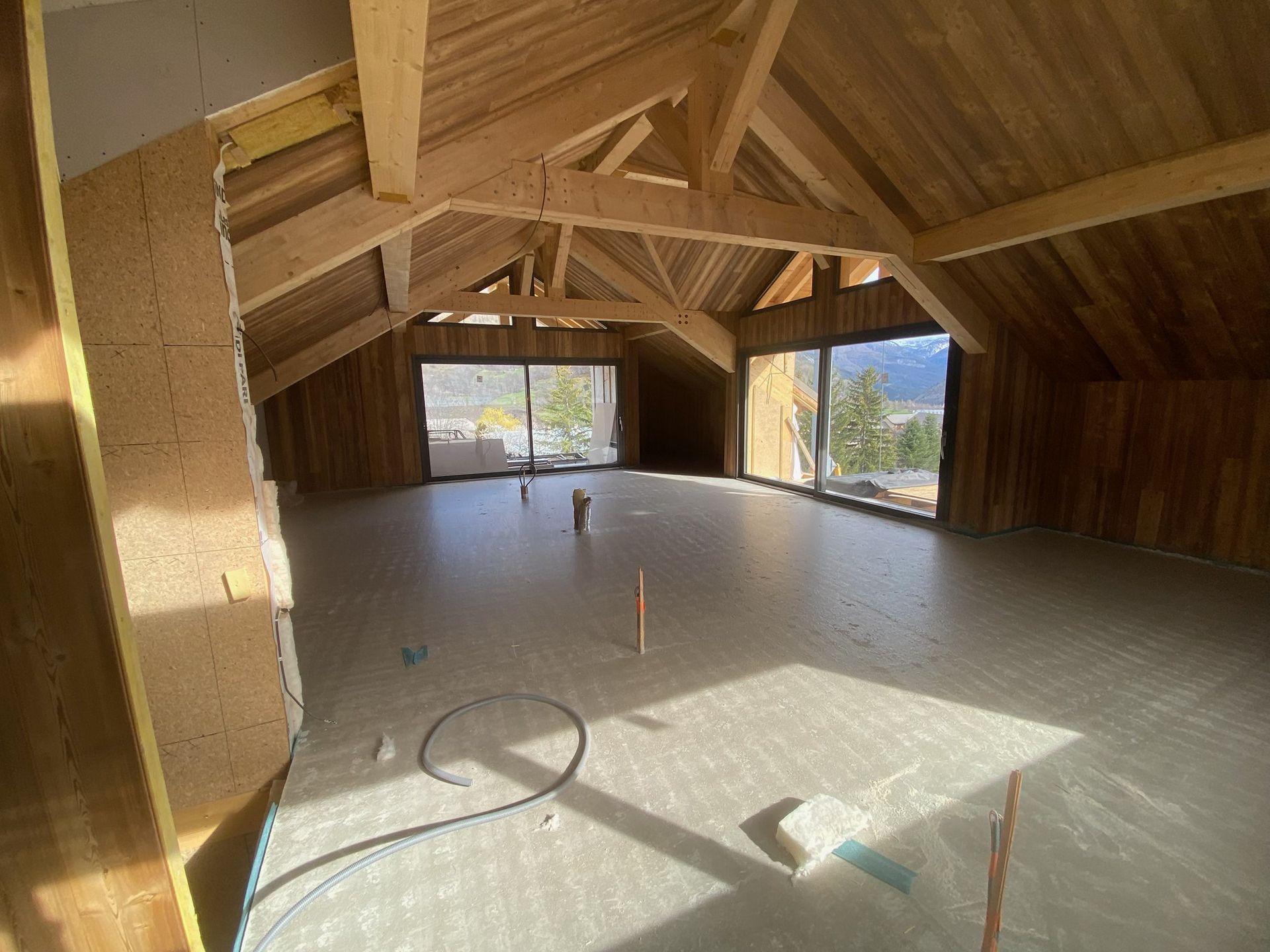 Une pièce mansardée avec des poutres en bois apparentes, un sol clair et de grandes fenêtres.