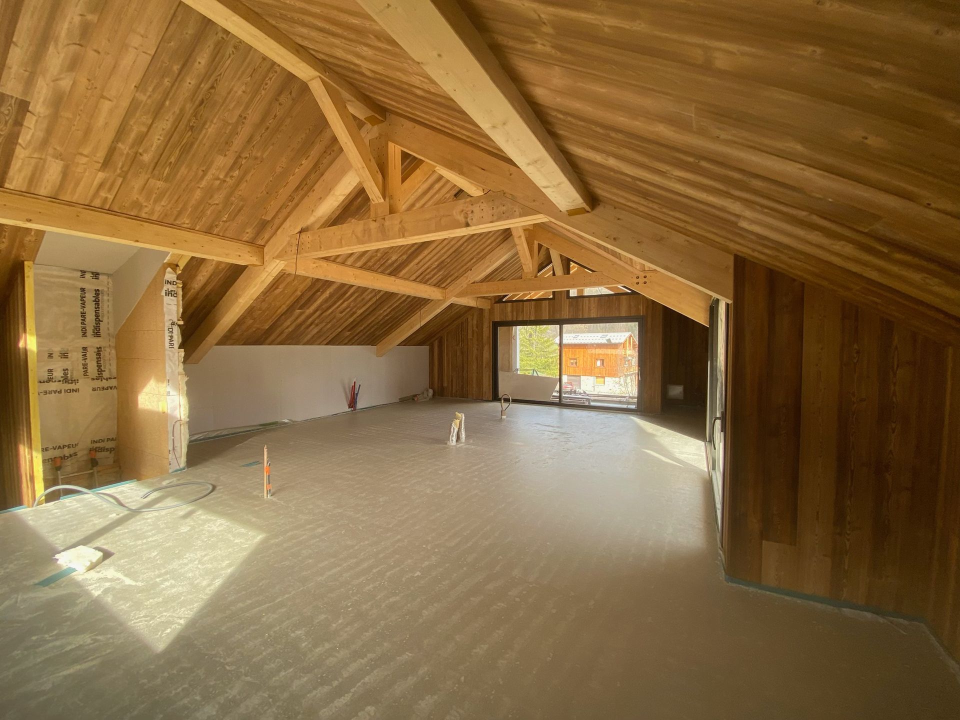 Une pièce mansardée inachevée avec charpente apparente et murs lambrissés.